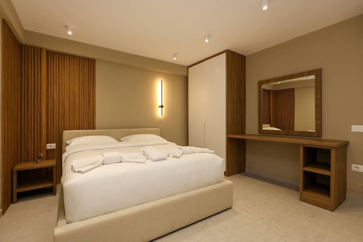 A modern bedroom with a large bed, bedside tables, a built-in bench, a mirror, and a wall-mounted light fixture.