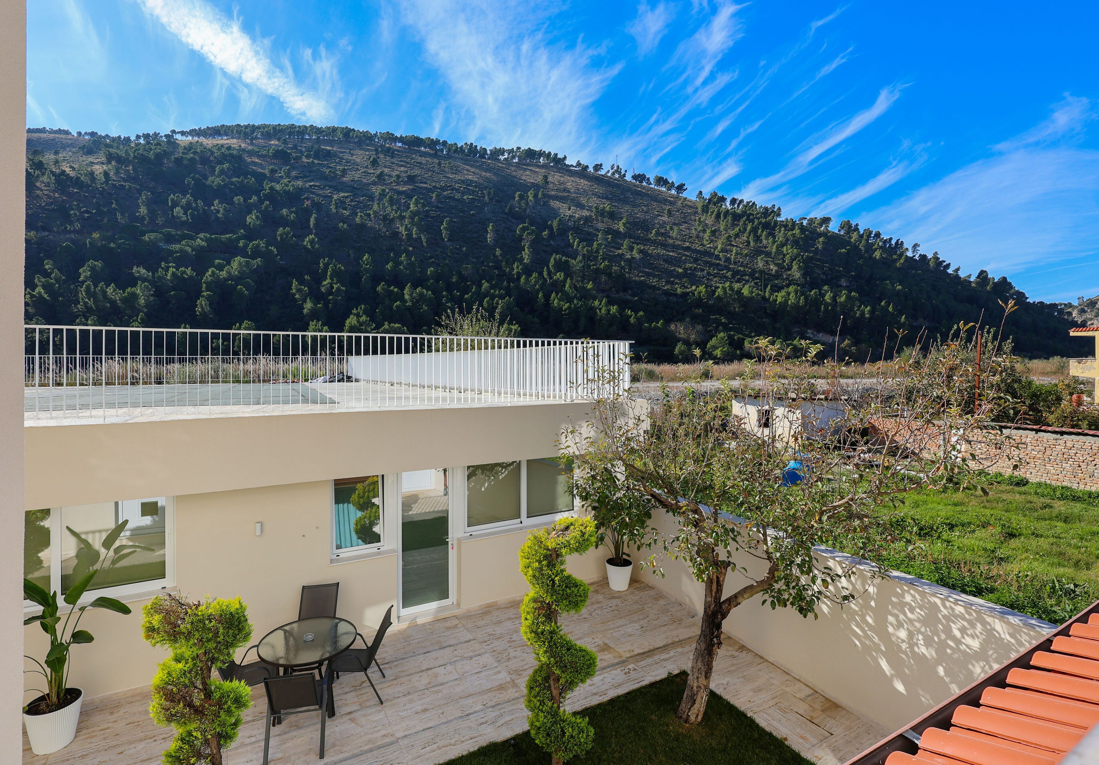 Uma casa moderna com dois andares, varanda, um pequeno pátio com mesa e cadeiras, e vista para uma colina arborizada.
