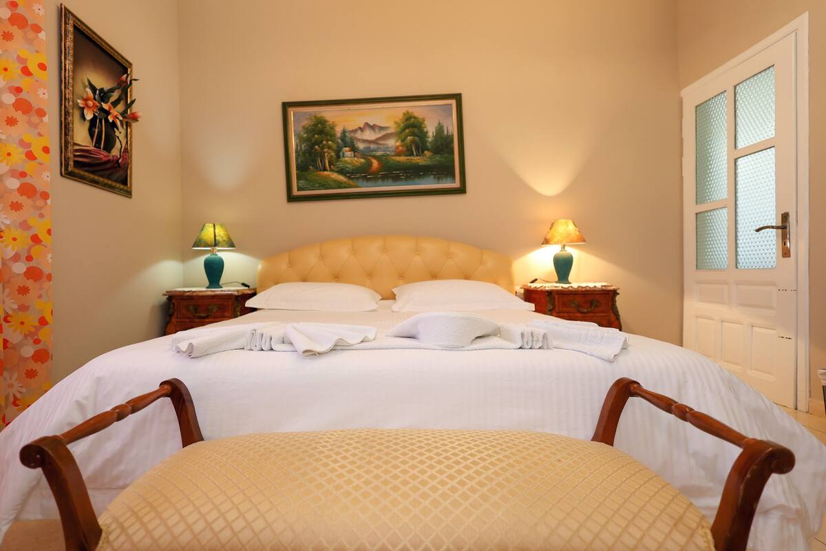 A neatly made bed with white linens, two bedside lamps, and a painting above the headboard.