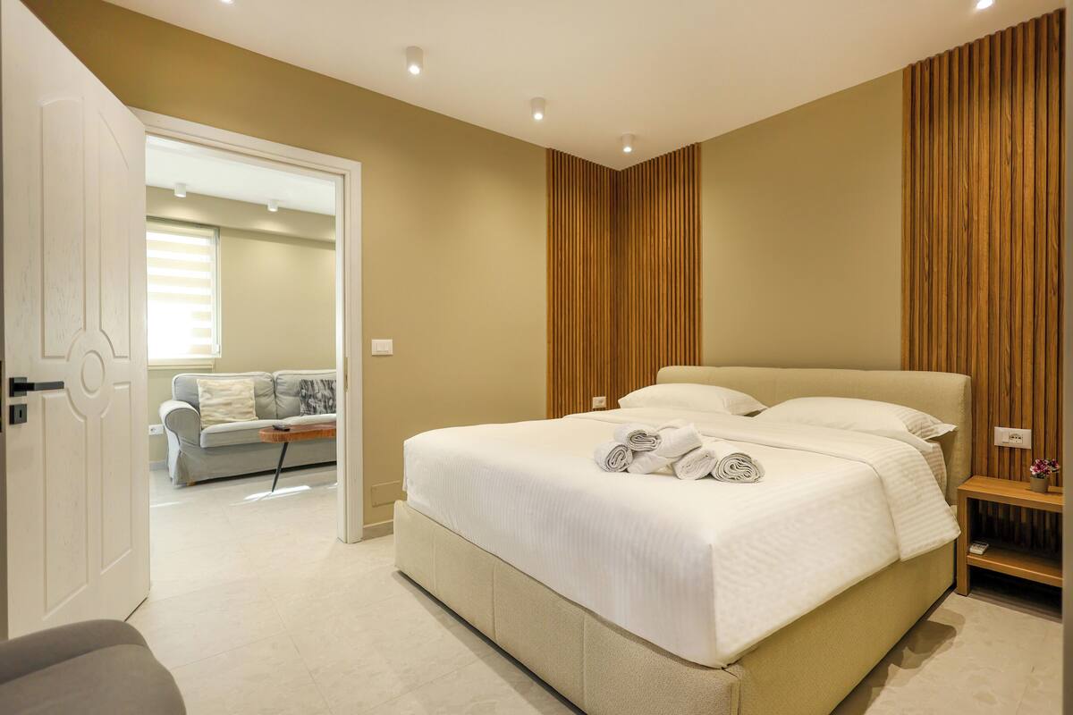 A modern bedroom with a large bed, a wooden panel wall, and a view into another room with a sofa.