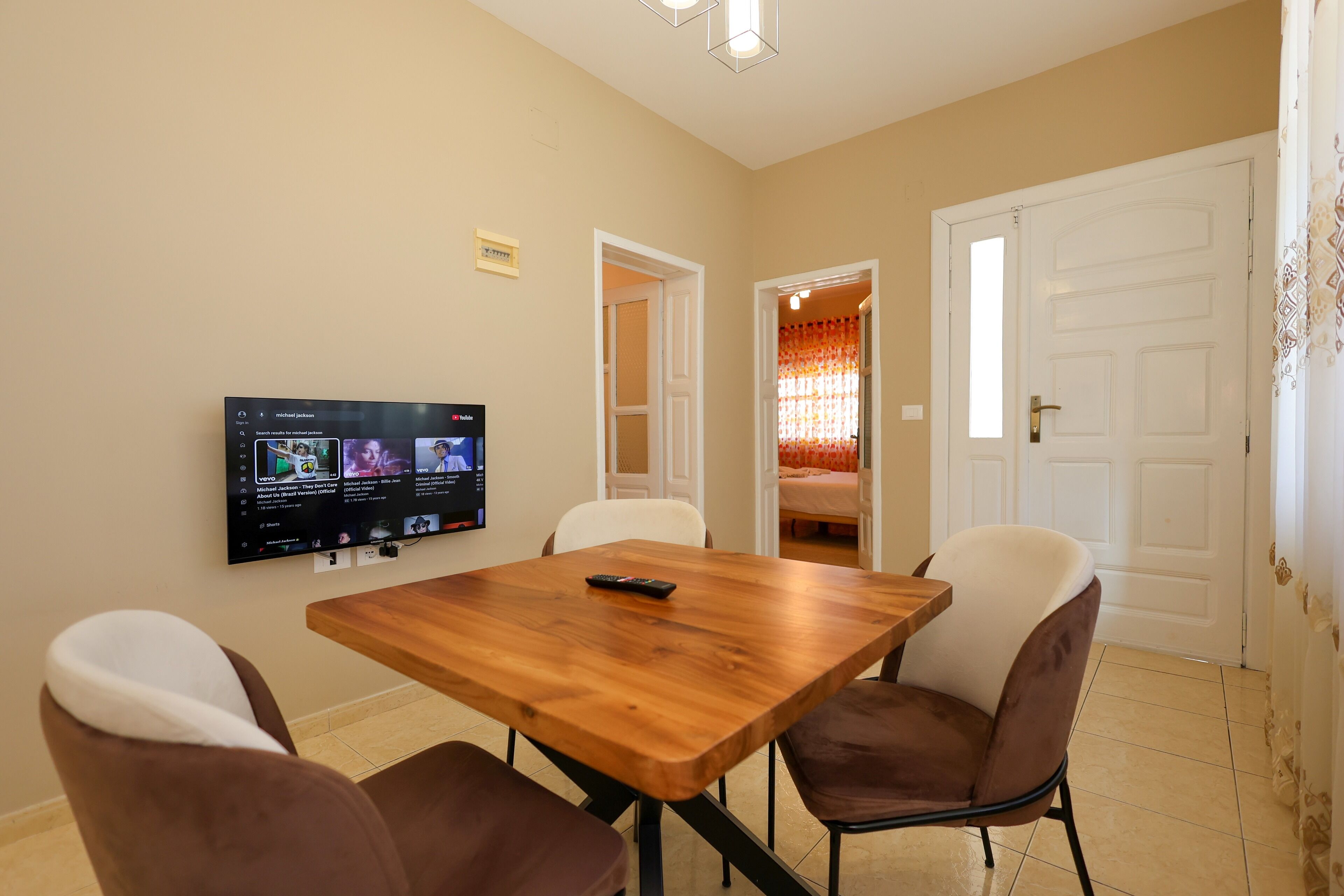 Un comedor con una mesa de madera y cuatro sillas, un televisor de pantalla plana en la pared, y una puerta que da a otra habitación.