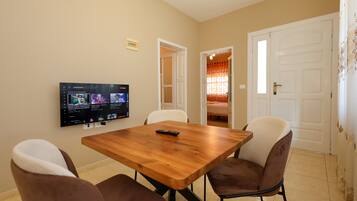 Un comedor con una mesa de madera y cuatro sillas, un televisor de pantalla plana en la pared, y una puerta que da a otra habitación.