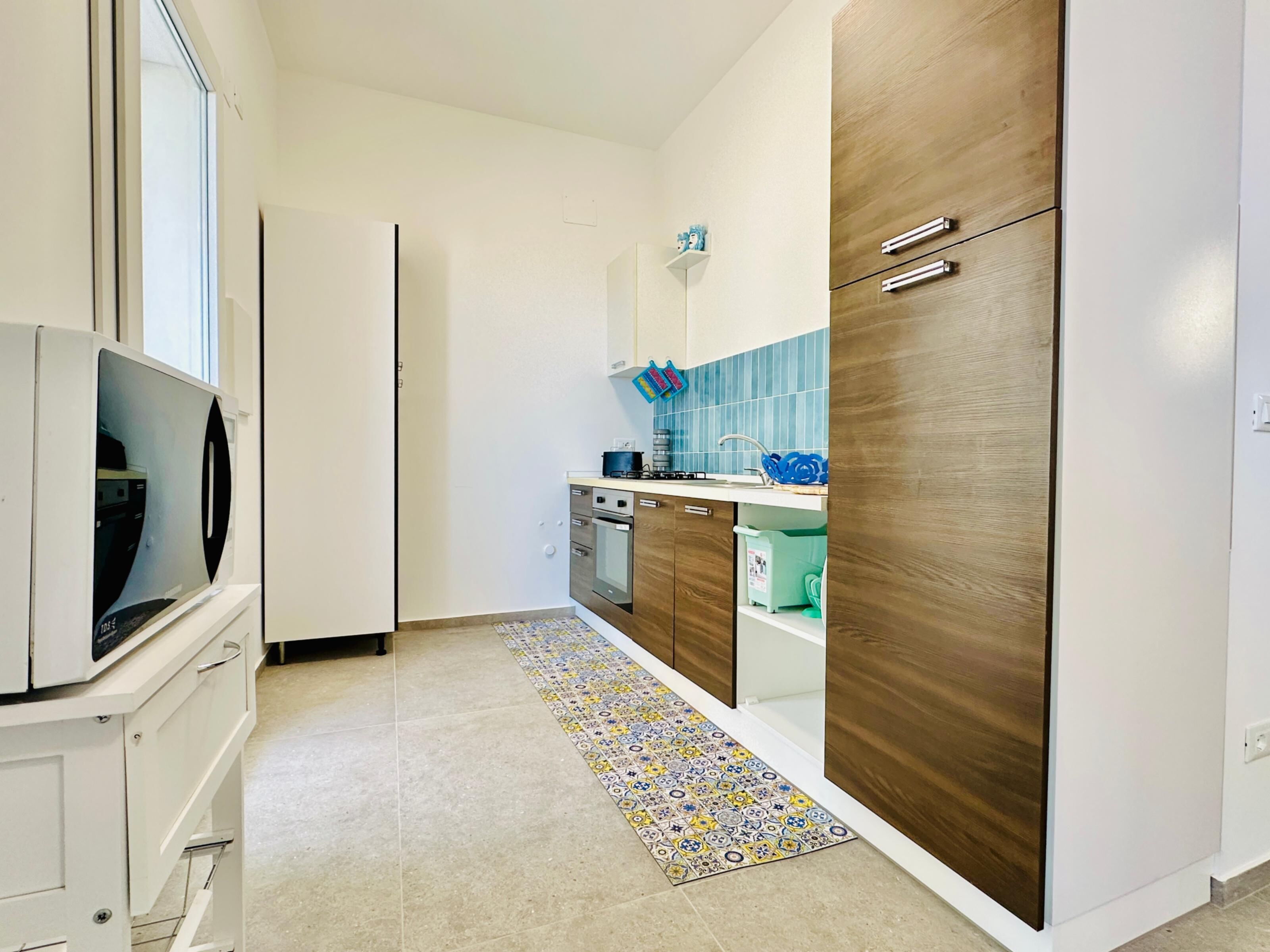Una cocina moderna con horno integrado, microondas y refrigerador. Hay una alfombra estampada en el suelo y un salpicadero azul sobre el fregadero.