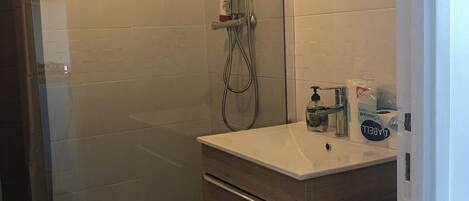 Une salle de bain moderne avec une douche à l’italienne en verre, un lavabo blanc avec un miroir au-dessus, et un meuble-lavabo en bois doté de tiroirs.