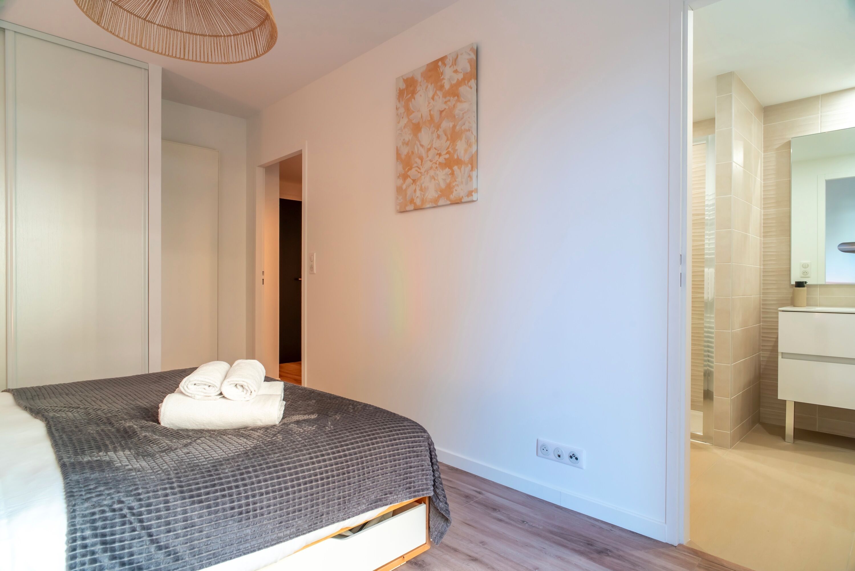 Une chambre à coucher avec un lit, une table de chevet, une œuvre d’art accrochée au mur et une salle de bain visible à travers une porte ouverte.