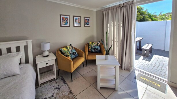 A hotel room with a bed, two armchairs, a small table, a bedside table, a lamp, and a view of the outside. - Ivy Place Studio Apartment (Cape Town)