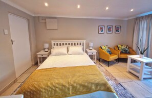Una habitación de hotel con una cama grande, dos mesitas de noche con lámparas, un sillón amarillo, una pequeña mesa blanca y una planta en maceta.