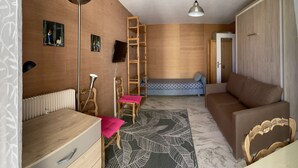 A compact room with a wooden floor, a sofa, a bed, a desk, a chair, a ladder, and a radiator.