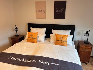 A neatly made bed with white bedding and two orange pillows, a black headboard, and two bedside tables with lamps. - Ferienhaus Niederrhein Kalkar - Traumhaus in Klein am Niederrhein (Kalkar)