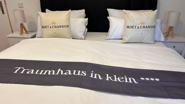 Un letto con biancheria bianca, due cuscini con la scritta 'MOËT & CHANDON CHAMPAGNE' e un corridore nero con la scritta 'Traumhaus in klein'.