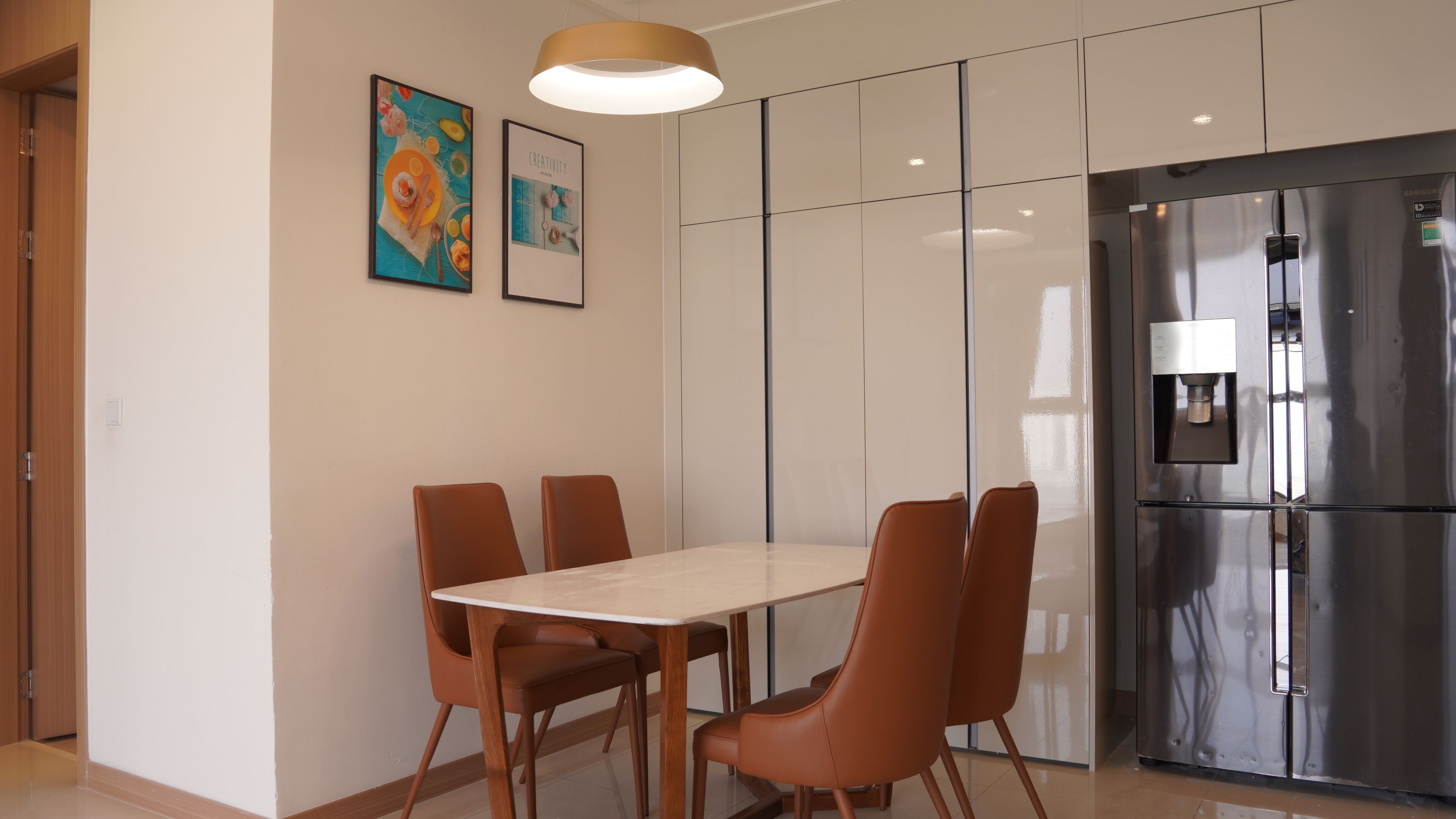A modern kitchen with a stainless steel refrigerator, a dining table with six chairs, and two framed artworks on the wall.