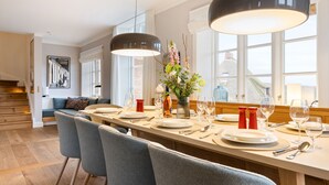Et spiseområde med et langt bord dækket til fire personer, en vase med blomster og udsigt til det frie landskab gennem store vinduer.