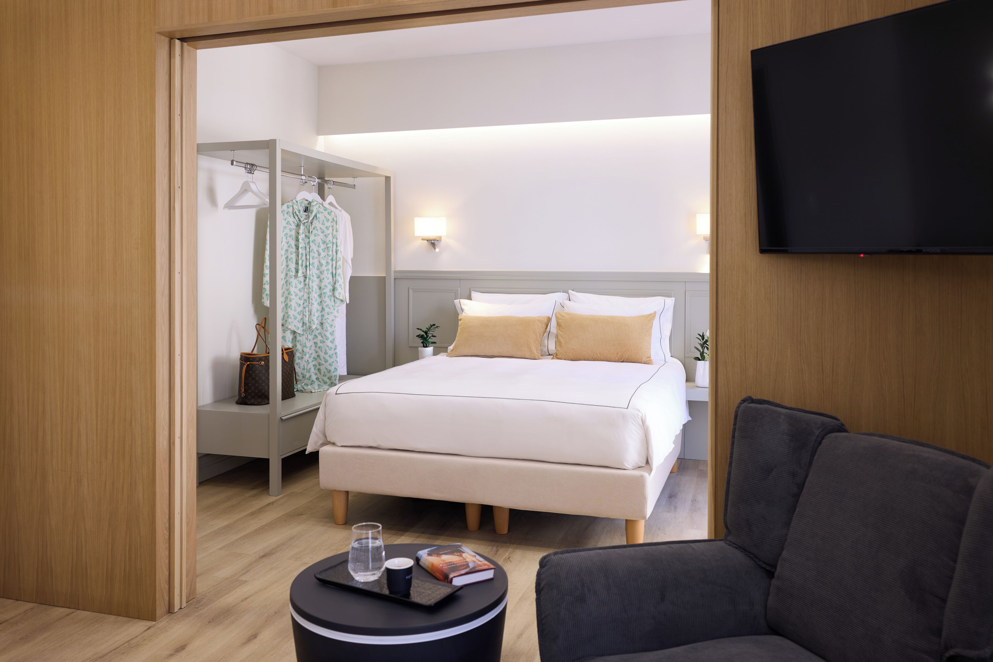 A modern hotel room with a bed, a TV, a seating area, and a walk-in closet.