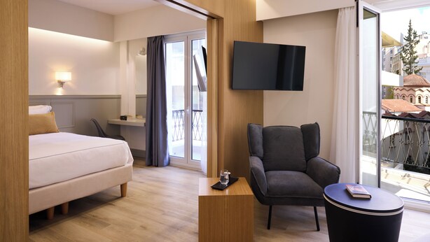 A hotel room with a bed, a chair, a small table, a TV, and a balcony with a view of buildings. - AthenSun Luxury Suites (Athens)