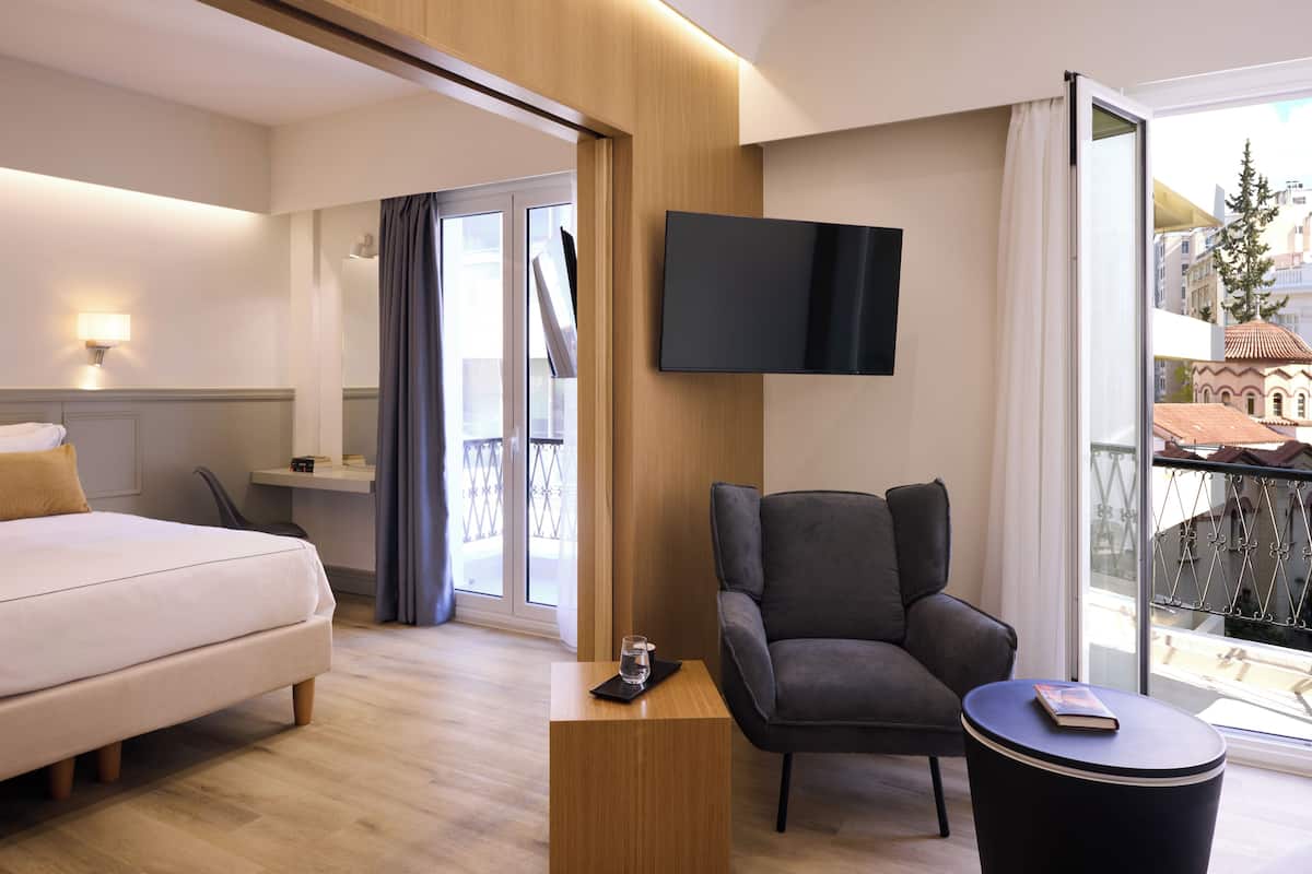 A hotel room with a bed, a chair, a small table, a TV, and a balcony with a view of buildings.