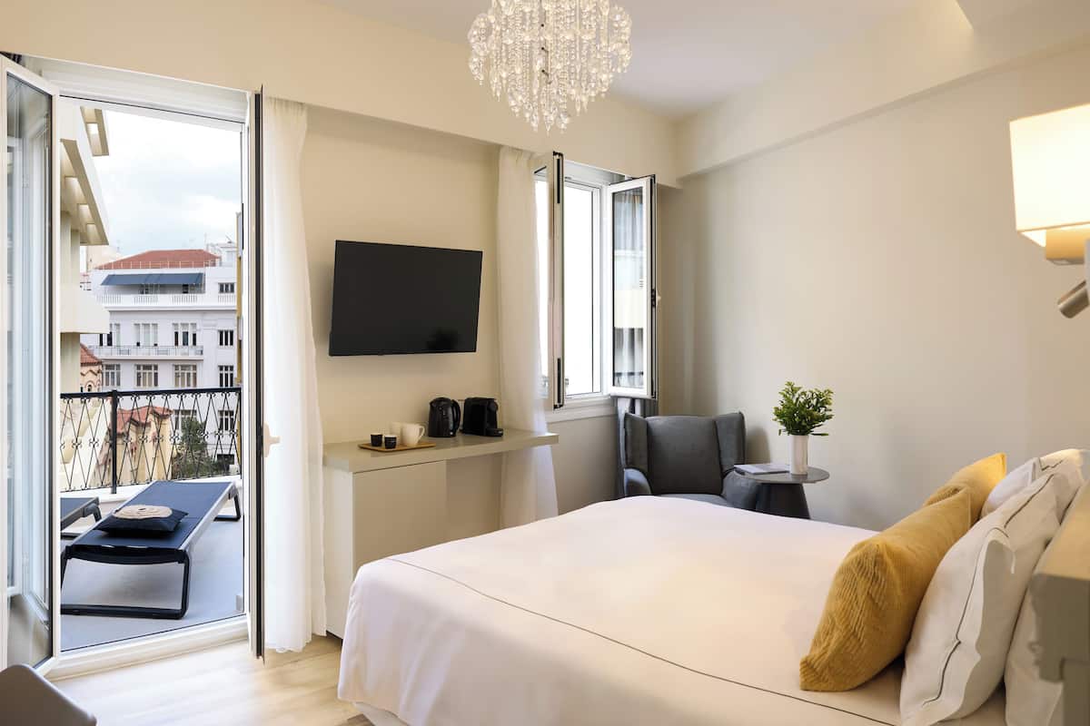 A hotel room with a bed, a desk with a coffee maker, a chair, a TV, and a balcony with a view of buildings.