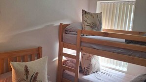Une chambre avec un lit superposé, comportant deux lits, chacun avec un oreiller et une couverture. Il y a une fenêtre avec des stores, et les murs de la chambre sont d’un blanc uni.