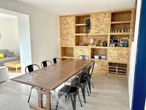 Uma área de jantar moderna com mesa e cadeiras de madeira, um aparador embutido com utensílios de cozinha e um sofá cinza ao fundo.