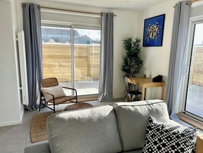 Uma sala de estar moderna com um sofá, uma mesa de jantar e uma cadeira. Há uma janela grande com cortinas, uma planta e um quadro na parede.