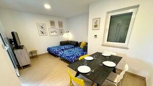 Un espacio habitable compacto con comedor, una cama con edredón azul estampado con flores, una pequeña mesa con sillas y un televisor sobre un mueble.