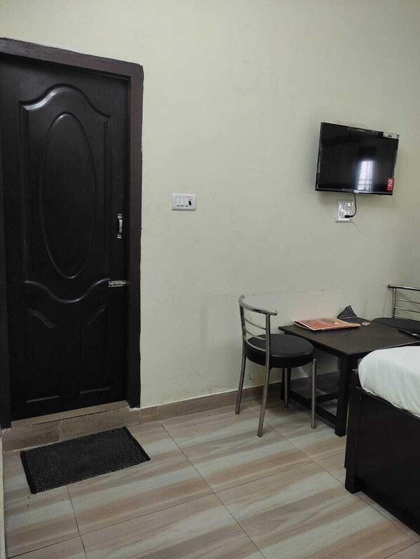 A small, simple room with a bed, a desk, a chair, a television mounted on the wall, and a door. - Wave Guest House (Kaladhungi)