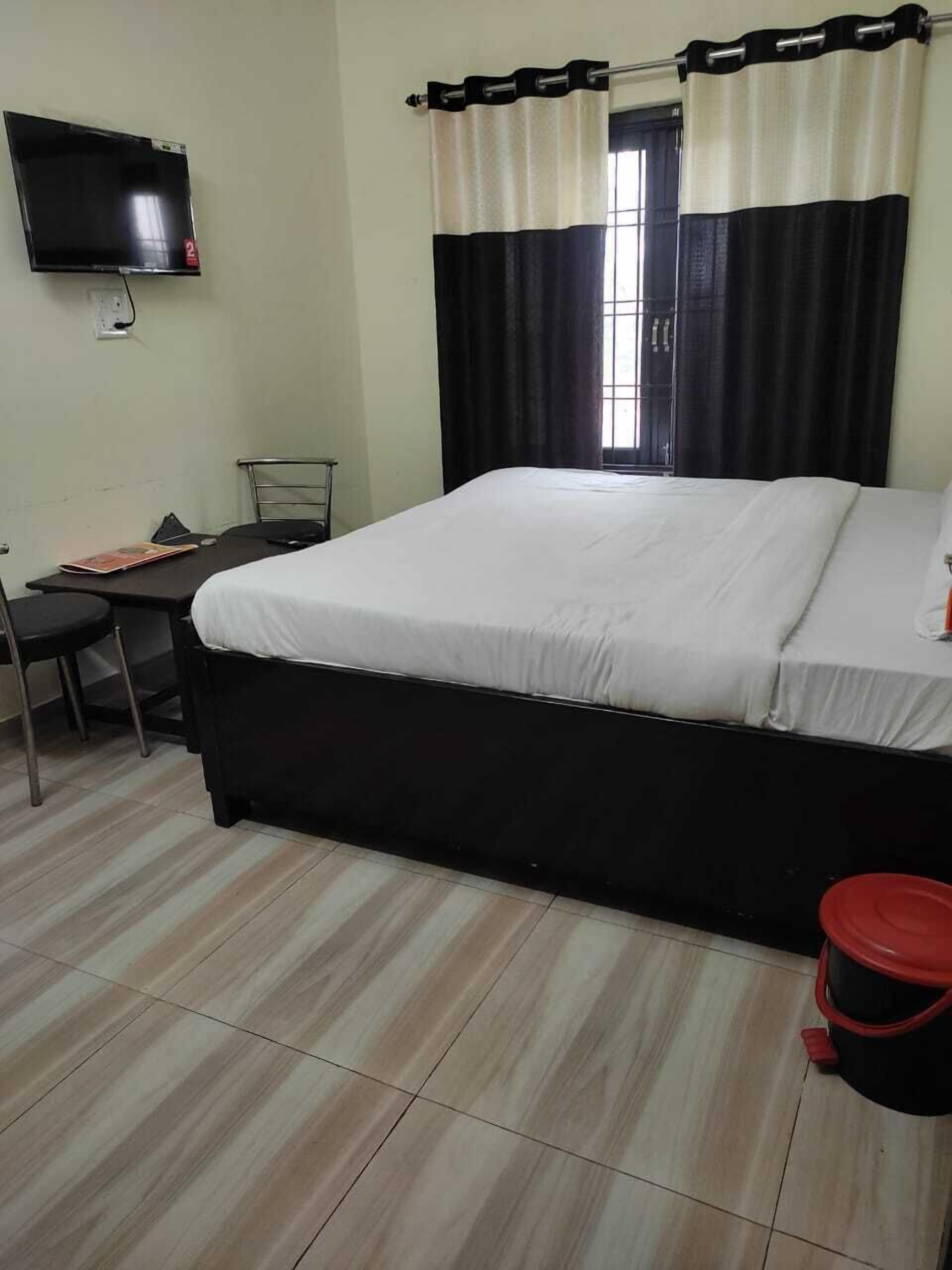 Een slaapkamer met een bed, een bureau, een stoel, een televisie en een raam met gordijnen.