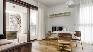 Ein modernes Wohnzimmer mit Sofa, einem Esstisch mit Stühlen, einem Fernseher und einem Balkon mit Blick auf Gebäude.