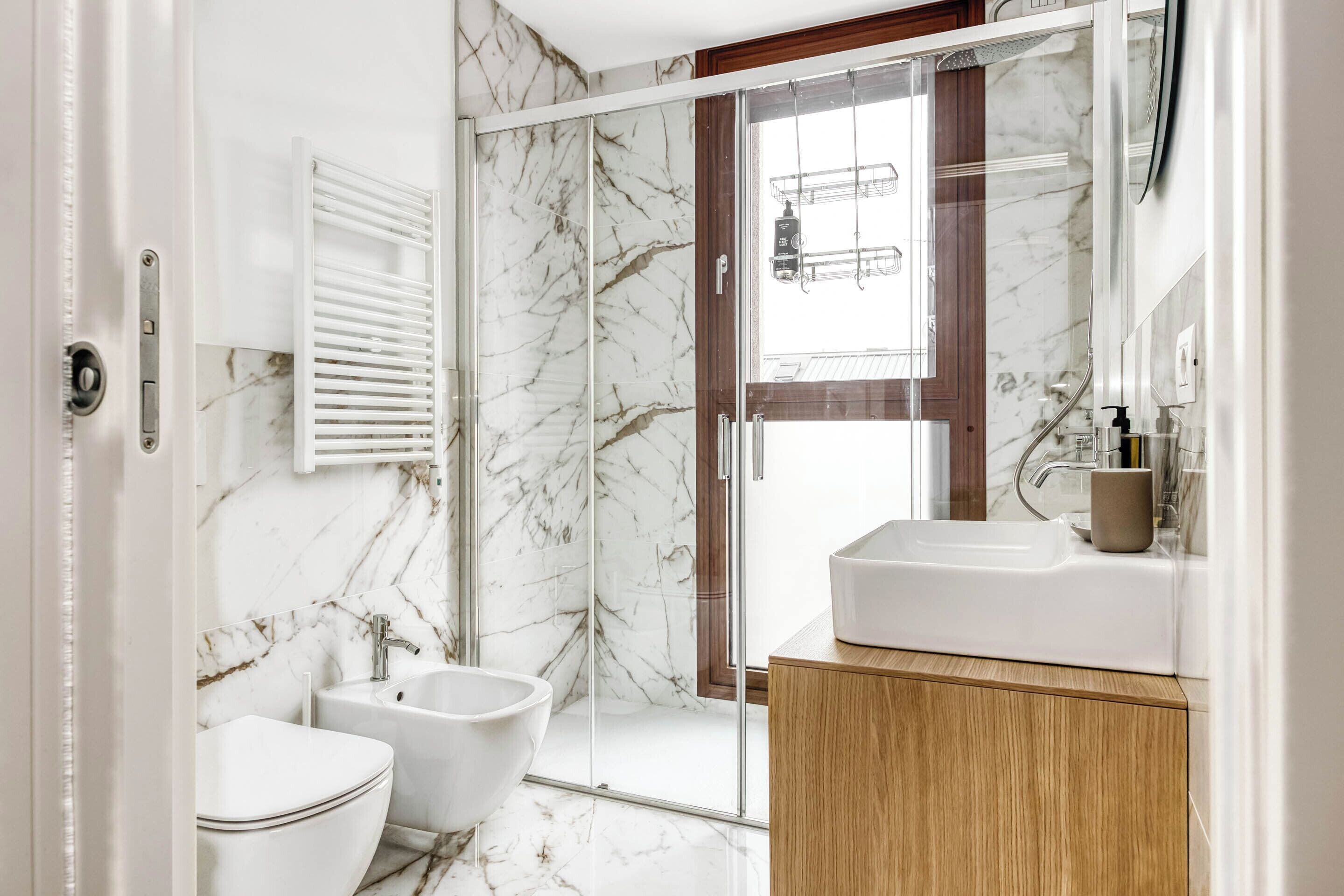 A modern bathroom with a marble floor, a glass shower enclosure, a white sink, and a wooden vanity.