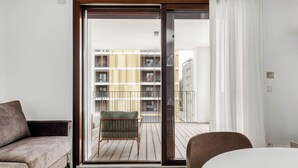 Ein modernes Zimmer mit Holzboden, einer Couch, einem Sessel und einem Tisch. Eine Glastür führt auf einen Balkon mit Blick auf Gebäude.