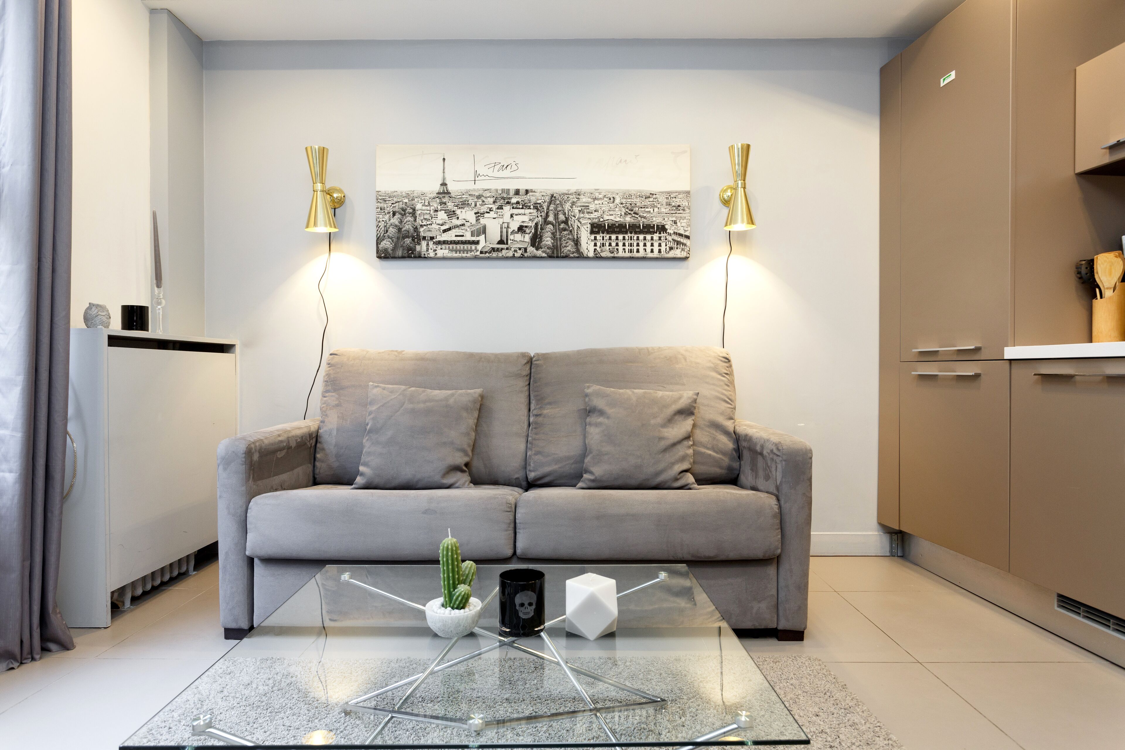 Un soggiorno moderno con un divano grigio, un tavolino in vetro con un cactus e una tazza a forma di teschio, e un quadro raffigurante un paesaggio urbano.
