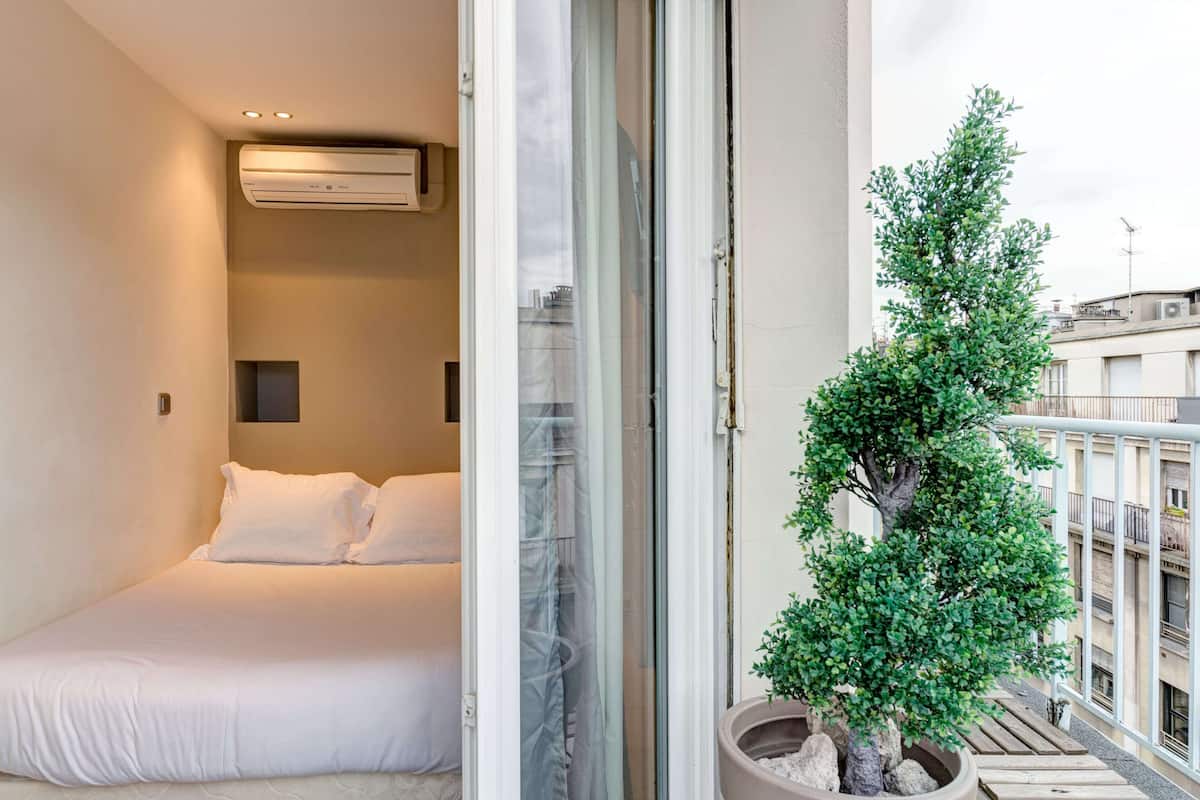 A bedroom with a bed, an air conditioning unit, and a view of a balcony with a potted tree.