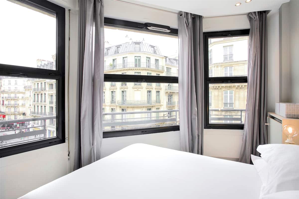 A hotel room with a large window, a bed, and a nightstand with a lamp.