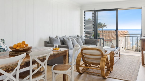Un salon moderne avec une table à manger en bois, des chaises blanches, un canapé avec des coussins gris et une vue sur la mer par une porte vitrée.
