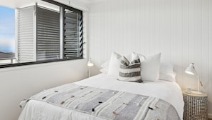 Une chambre à coucher avec un lit, des oreillers, une lampe de chevet, une fenêtre avec des stores et une vue sur la mer.