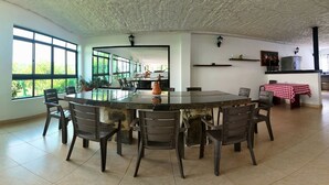 Un comedor amplio con una gran mesa redonda rodeada de sillas, una cocina con refrigerador y ventanas grandes que ofrecen vistas a zonas verdes.