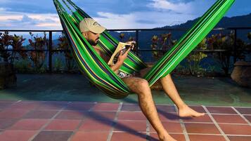 Una persona descansando en una hamaca en un patio con baldosas, con vistas a montañas y plantas.