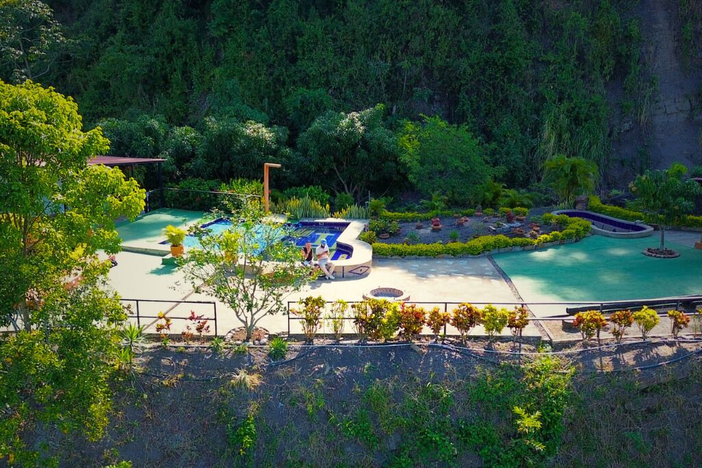 Un área de piscina con un elemento acuático circular, rodeada de abundante vegetación y un risco rocoso.