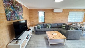 Un salon doté d’un téléviseur à écran plat, d’un canapé gris agrémenté de coussins, d’une table basse en bois et d’un tableau abstrait aux couleurs vives accroché au mur.