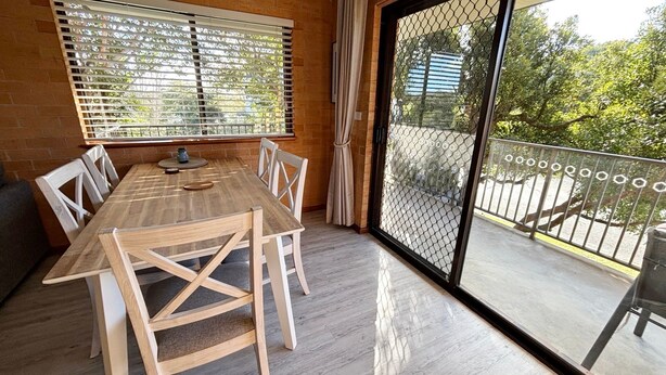 Un coin repas avec une table et des chaises en bois, un mur en briques et une porte-fenêtre coulissante donnant sur un balcon agrémenté d’une rambarde en métal.