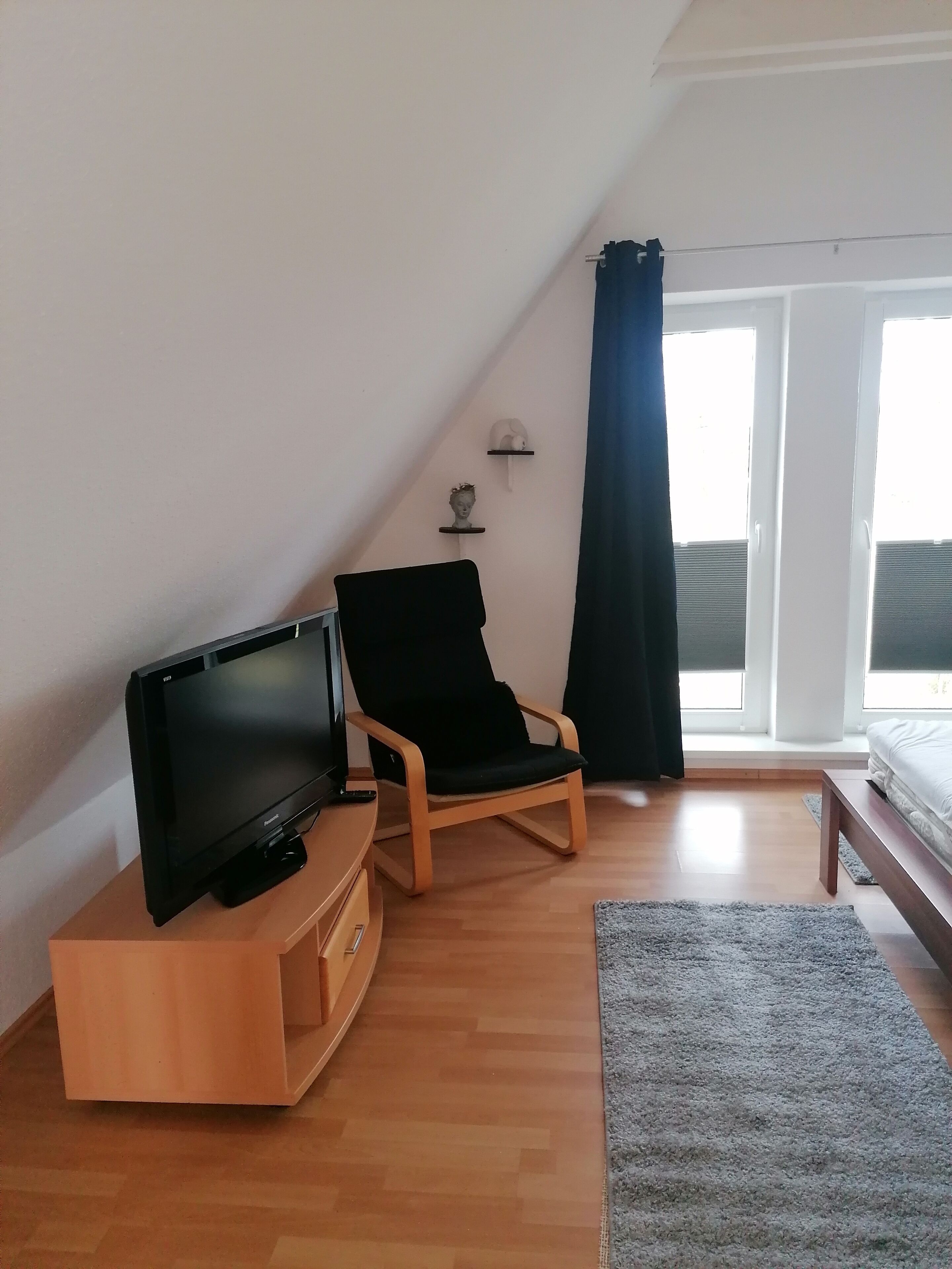 Ein Zimmer mit einem Flachbildfernseher auf einem Holzständer, einem schwarzen gepolsterten Sessel, einer Holzbank und einem grauen Teppich.