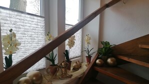 Una escala amb barana de fusta, una finestra amb persianes blanques, i diversos objectes decoratius com ara plantes en testos, una estàtua de Buda i esfères daurades.