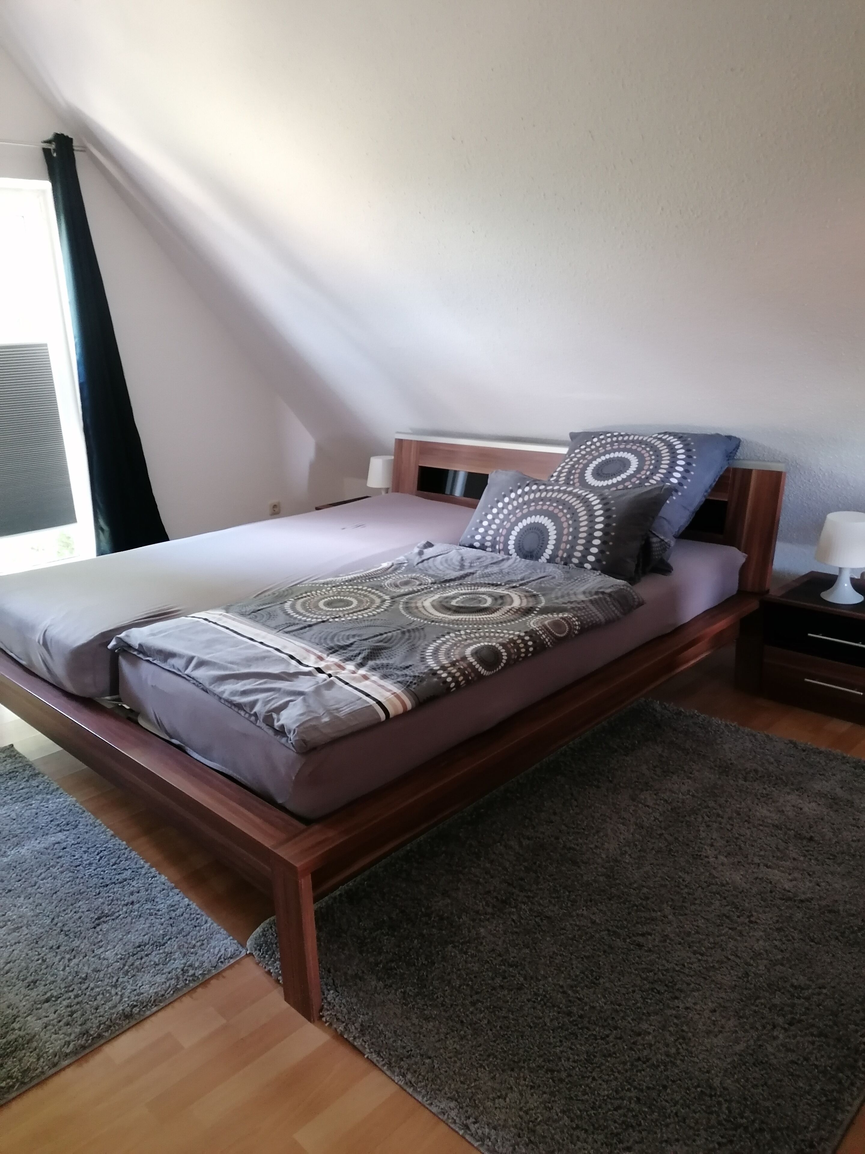 Una cama individual con un edredón estampado y almohadas, una mesita de noche con una lámpara, y una ventana con cortinas.