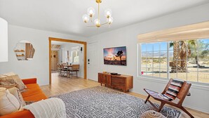 A spacious living room with a large window, a wooden floor, a modern chandelier, and a cozy orange sofa. - Desert escape w/ hot tub, pool, fire pit & fully fenced yard — 15 min to JTNP. Perfect for dog-friendly getaways. 🌵✨ (Joshua Tree)