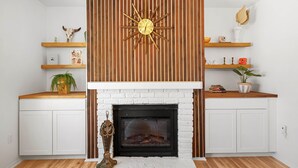 A modern living room with a fireplace, wooden panel wall, and shelves with decorative items. - Desert escape w/ hot tub, pool, fire pit & fully fenced yard — 15 min to JTNP. Perfect for dog-friendly getaways. 🌵✨ (Joshua Tree)