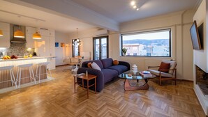 A modern living room with a sofa, coffee table, and bar stools. A kitchen with a large window and city view is visible in the background. - Athens Panorama Penthouse by the Acropolis (Athina)