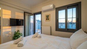 A hotel room with a bed, a TV, a plant, and a view of the city through the window. - Athens Panorama Penthouse by the Acropolis (Athina)