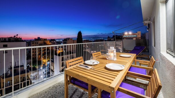 A balcony with a wooden table set for dining, purple cushions, and a view of the cityscape at dusk. - Athens Panorama Penthouse by the Acropolis (Athina)