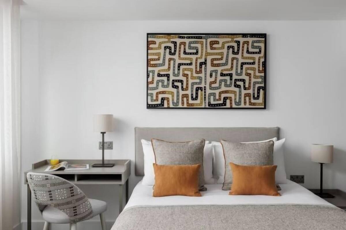 Habitación de hotel ordenada con una cama, mesitas de noche, una silla y una obra de arte colgada en la pared.