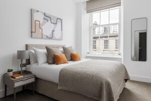 Une chambre à coucher comprenant un lit, une table de chevet, une lampe, une petite plante et une fenêtre donnant sur des immeubles.
