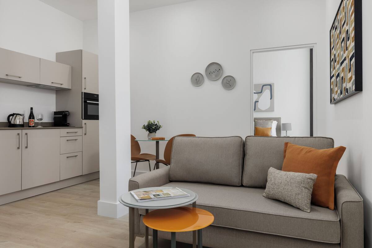 A modern living room with a grey sofa, a small round table, and a kitchen area with cabinets and appliances.